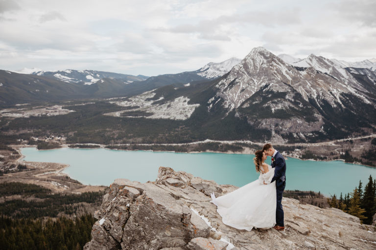 A couple on their mountain hiking elopement in Kananaskis, Alberta, Canadian Rockies. Captured by Banff wedding and elopement photographers. The couples are standing on the edge of a cliff surrounded by mountain range with turquoise coloured alpine lake behind them on a windy sunny day.