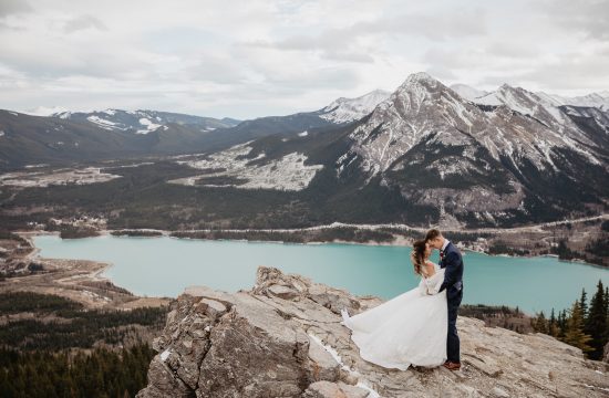 A couple on their mountain hiking elopement in Kananaskis, Alberta, Canadian Rockies. Captured by Banff wedding and elopement photographers. The couples are standing on the edge of a cliff surrounded by mountain range with turquoise coloured alpine lake behind them on a windy sunny day.