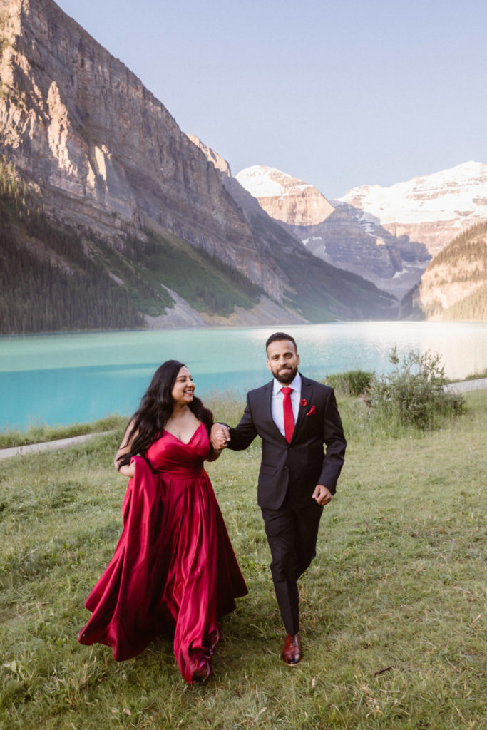 An engaged couple running together at Lake Louise. She is wearing a a red dress and he is wearing a suit with red tie
