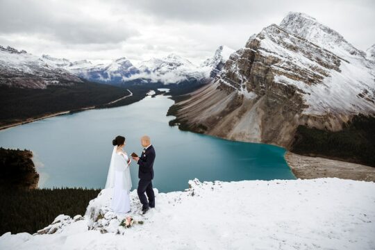 A couple getting married on top of a mountain, at the edge of a cliff overlooking a blue lake.