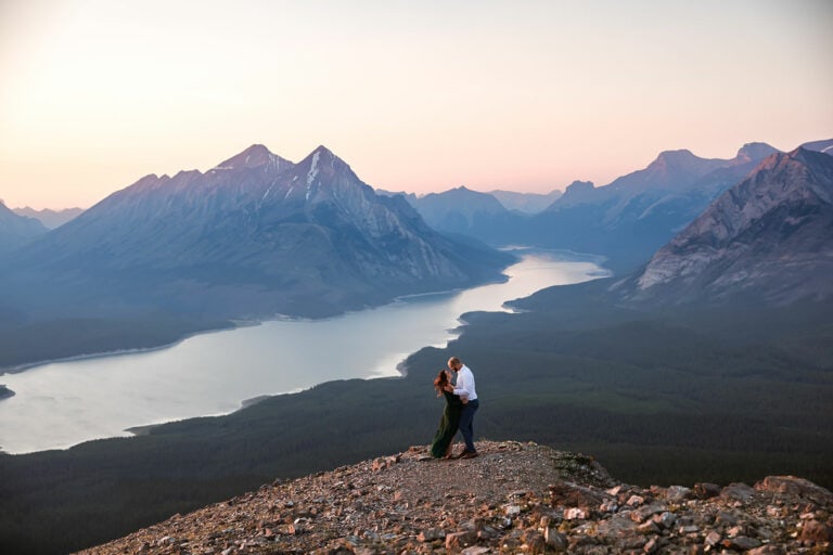 Engaged couples looking at each other while standing on top of a mountain during sunset