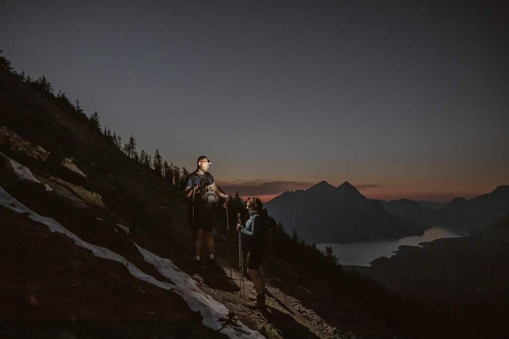 an engaged couples hiking down the mountain in the dark while carrying their pup after their engagement session in banff.