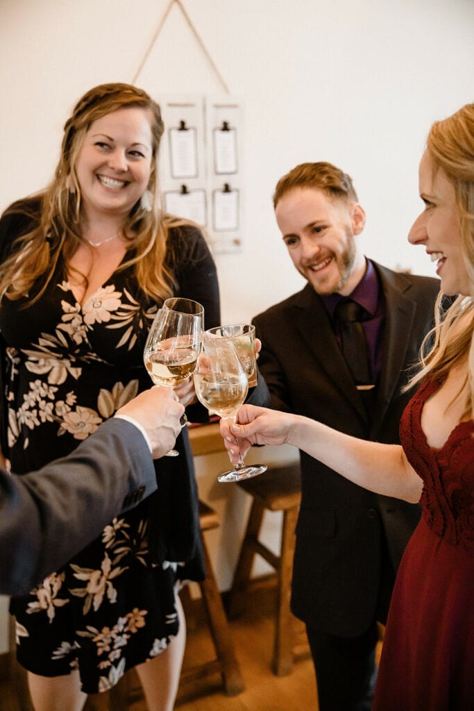 Three smiling people in wedding attire clinking wine glasses together with a fourth person just out of frame. 