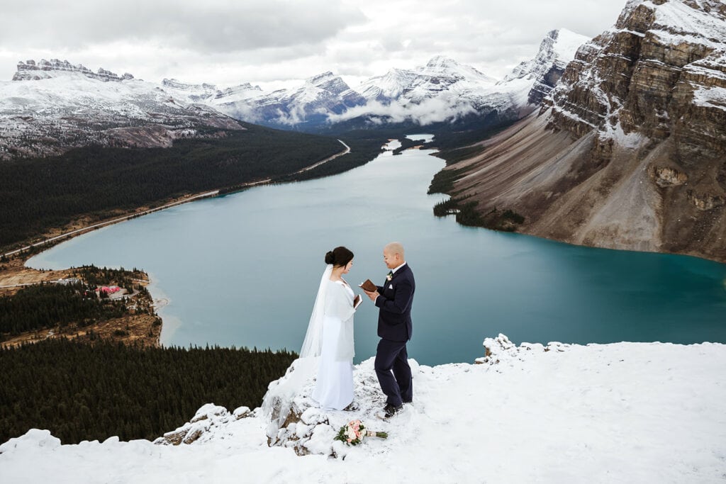 A bride in a white dress and a groom in a black suit exchanging private vows on a snowy ledge overlooking bow lake, with snow covered mountains in the background