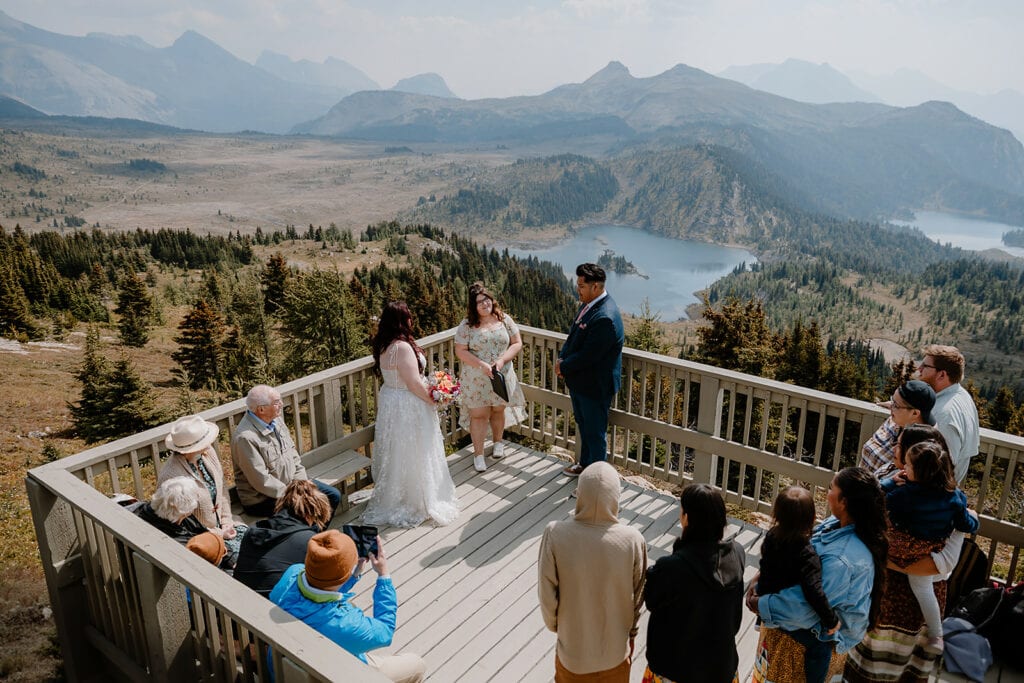 A bride and groom surrounded by their friends and family, all standing on a wooden platform overlooking an alpine valley and lake below, mountains in the background