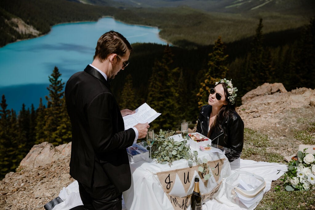 A groom wearing a black suit reading letters that his family and friends wrote to them for their elopement. The bride is listening with a smile on her face. 