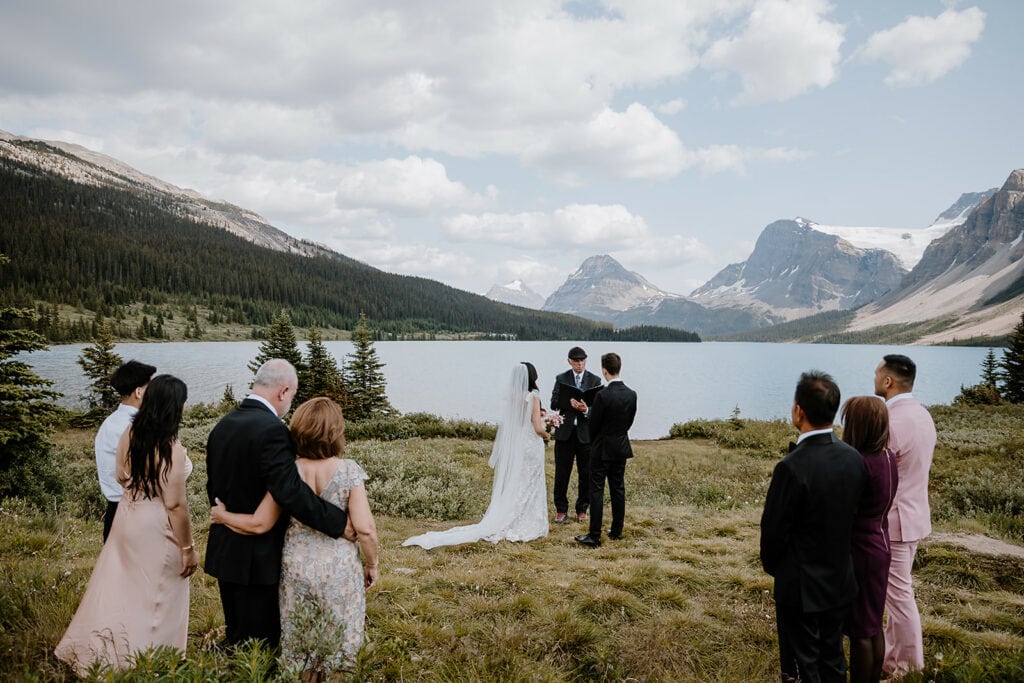 A couple exchanging vows in front of an officiant, mountains and lake in background, small group of friends and family in foreground