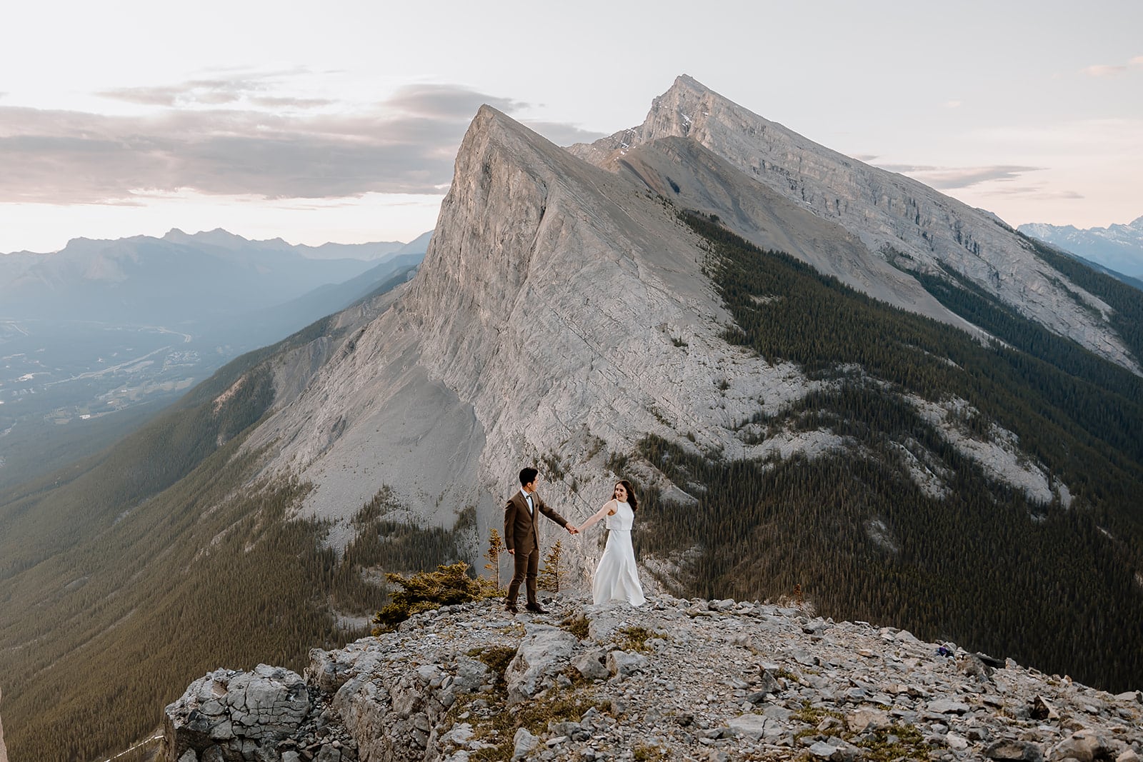 A BRIDE AND GROOM HOLDING HANDS ON TOP OF A MOUNTAIN