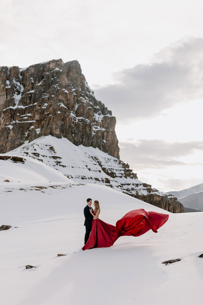 Bride and groom to be standing on a snowy mountain top on their photoshoot in Banff. she wearing a red dress with long flowy train and he's wearing a suit