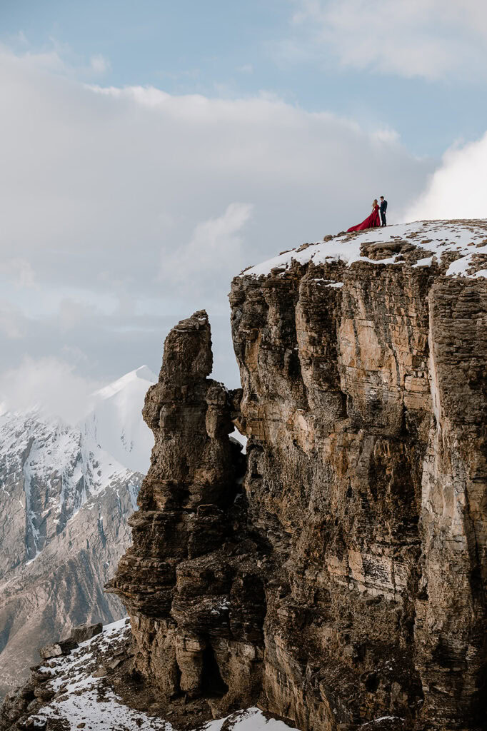 Bride and groom to be standing on the side of a cliff over looking the snowy mountain tops on their photoshoot in Banff. she wearing a red dress with long flowy train and he's wearing a suit