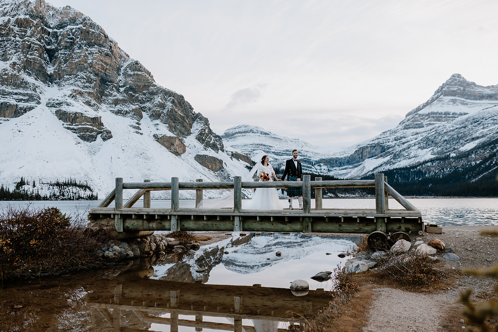 a bride and groom walk across a bridge with snowy mountains in the background 
