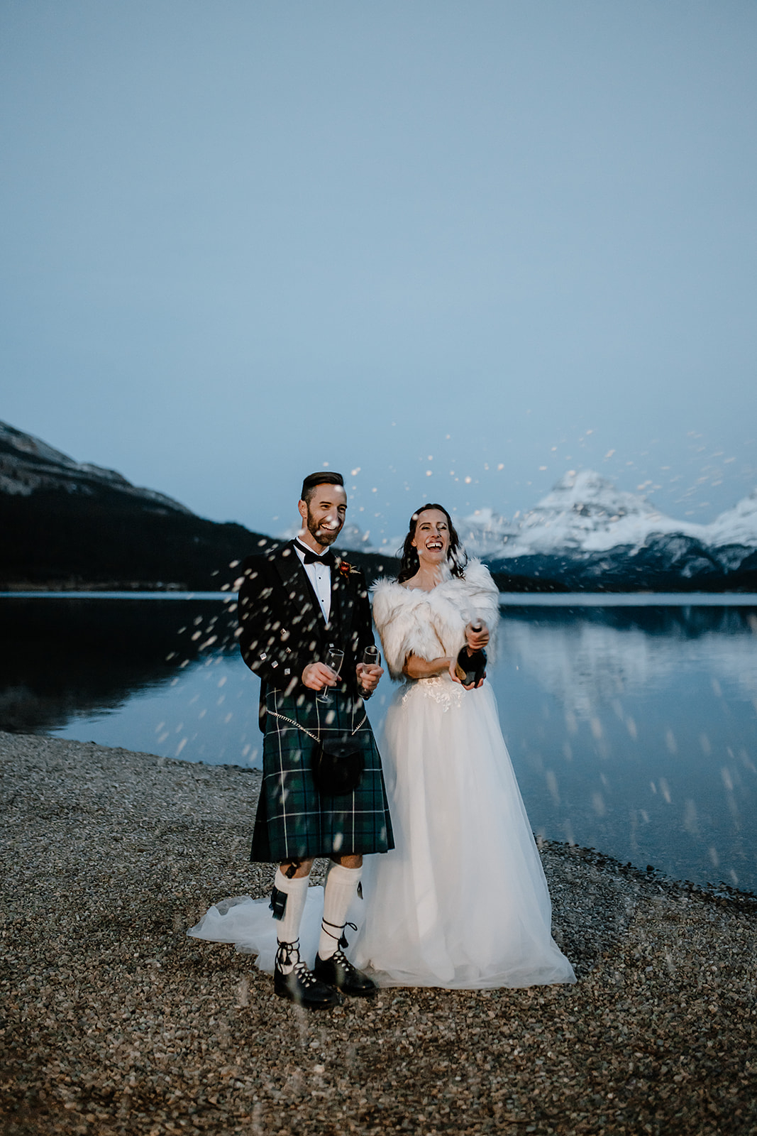 a couple stands on the shore of their Bow Lake wedding while popping a bottle of champagne to celebrate 