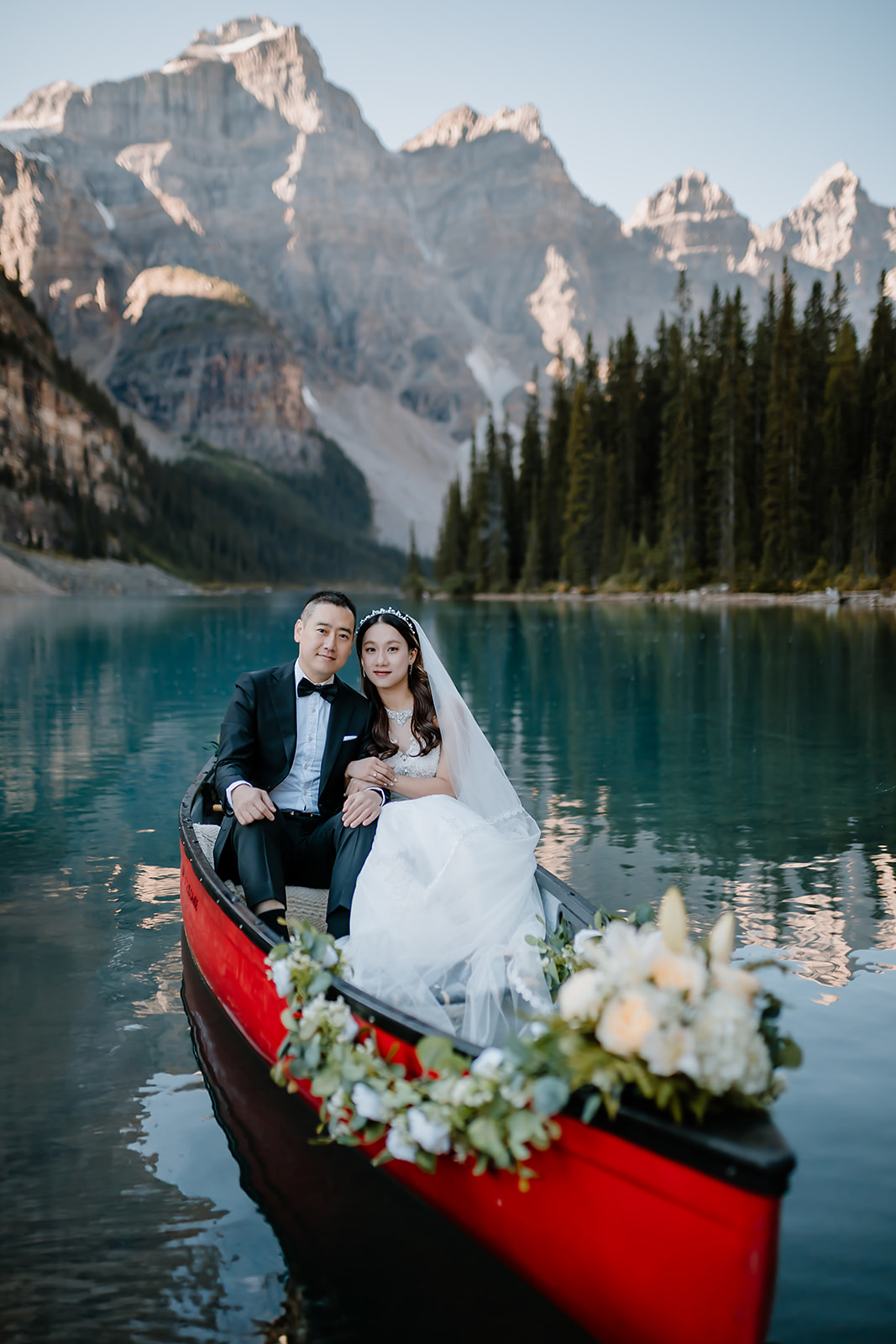 a bride and groom sit in a red canoe decorated with flowers during their pre-wedding canoe ride photoshoot at Moraine Lake 