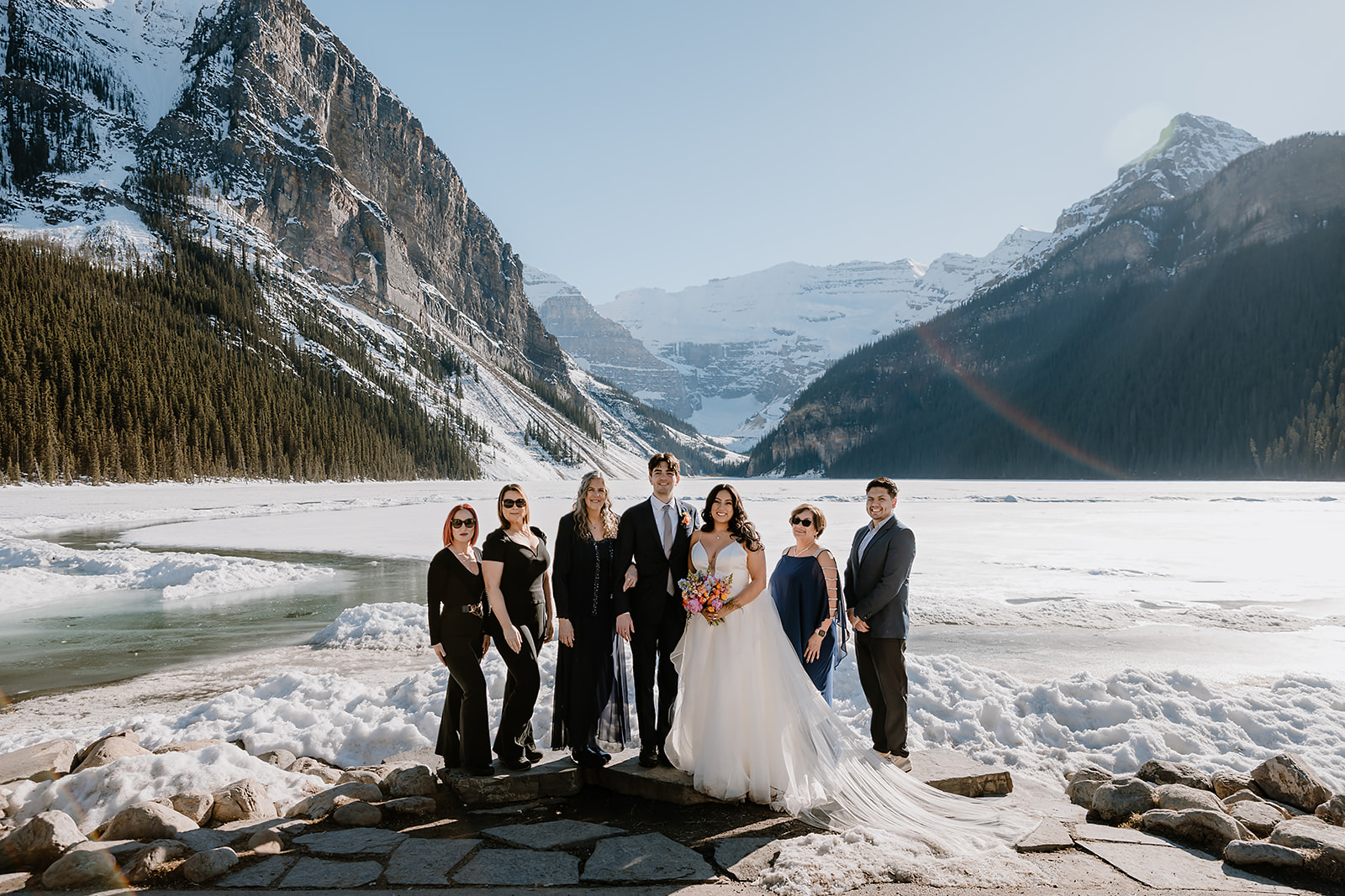 get-married-in-Banff a small wedding party gathers by a frozen lake with mountains in the background- get married in Banff