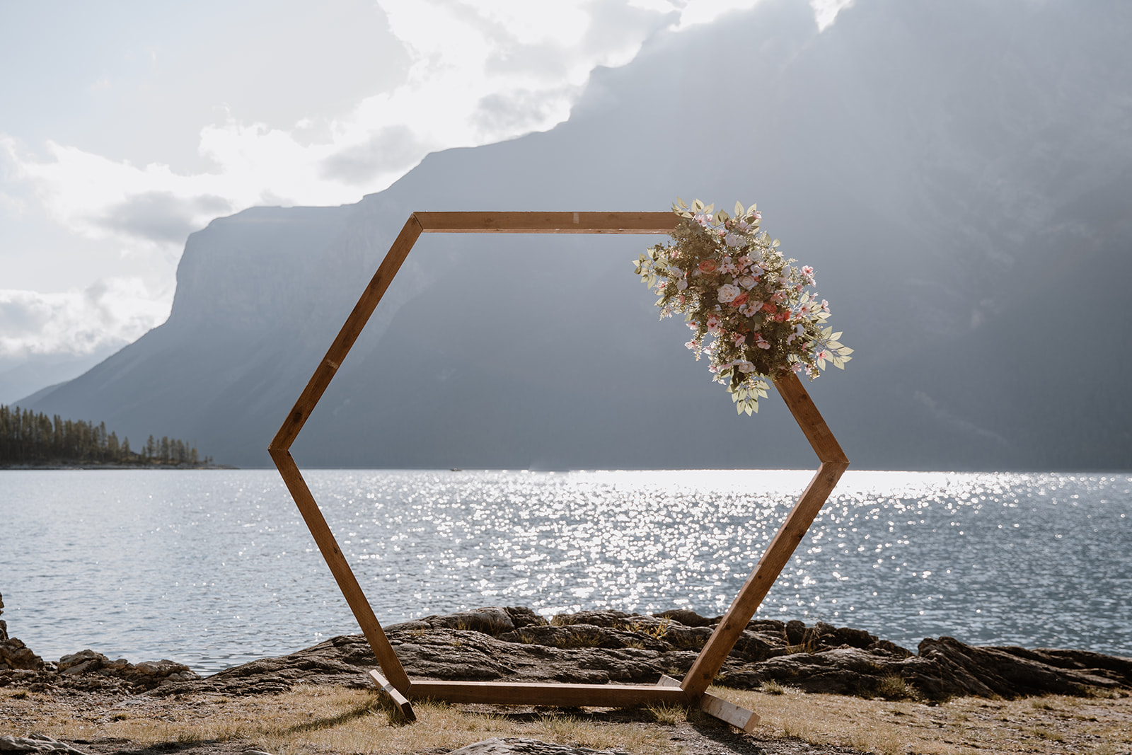 lake-Minnewanka-wedding A wooden hexagon arch adorned with flowers stands beside a shimmering lake and dramatic mountains.