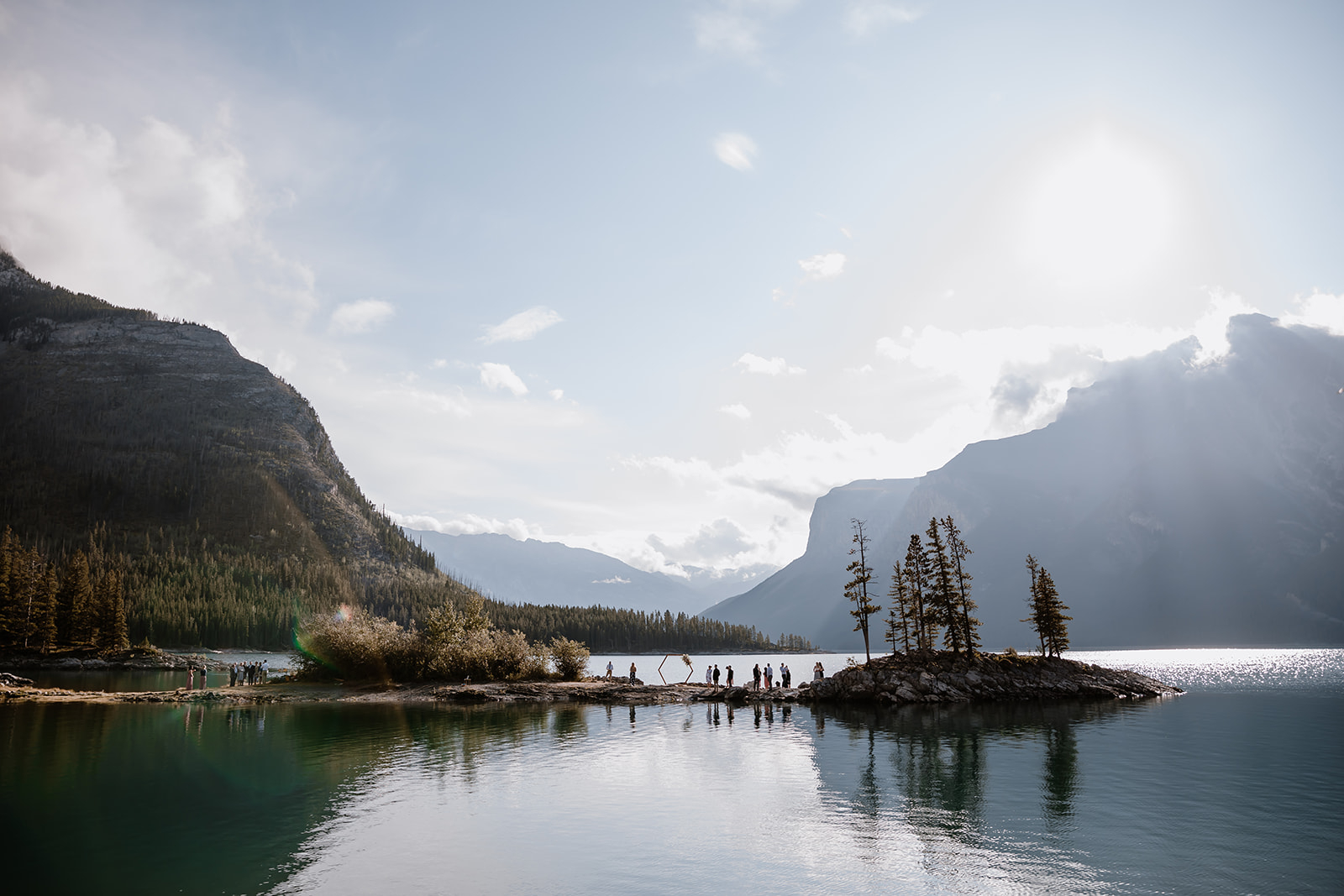lake-Minnewanka-wedding A small wedding gathers lakeside under sunlight, surrounded by mountains, trees, and shimmering water reflections.