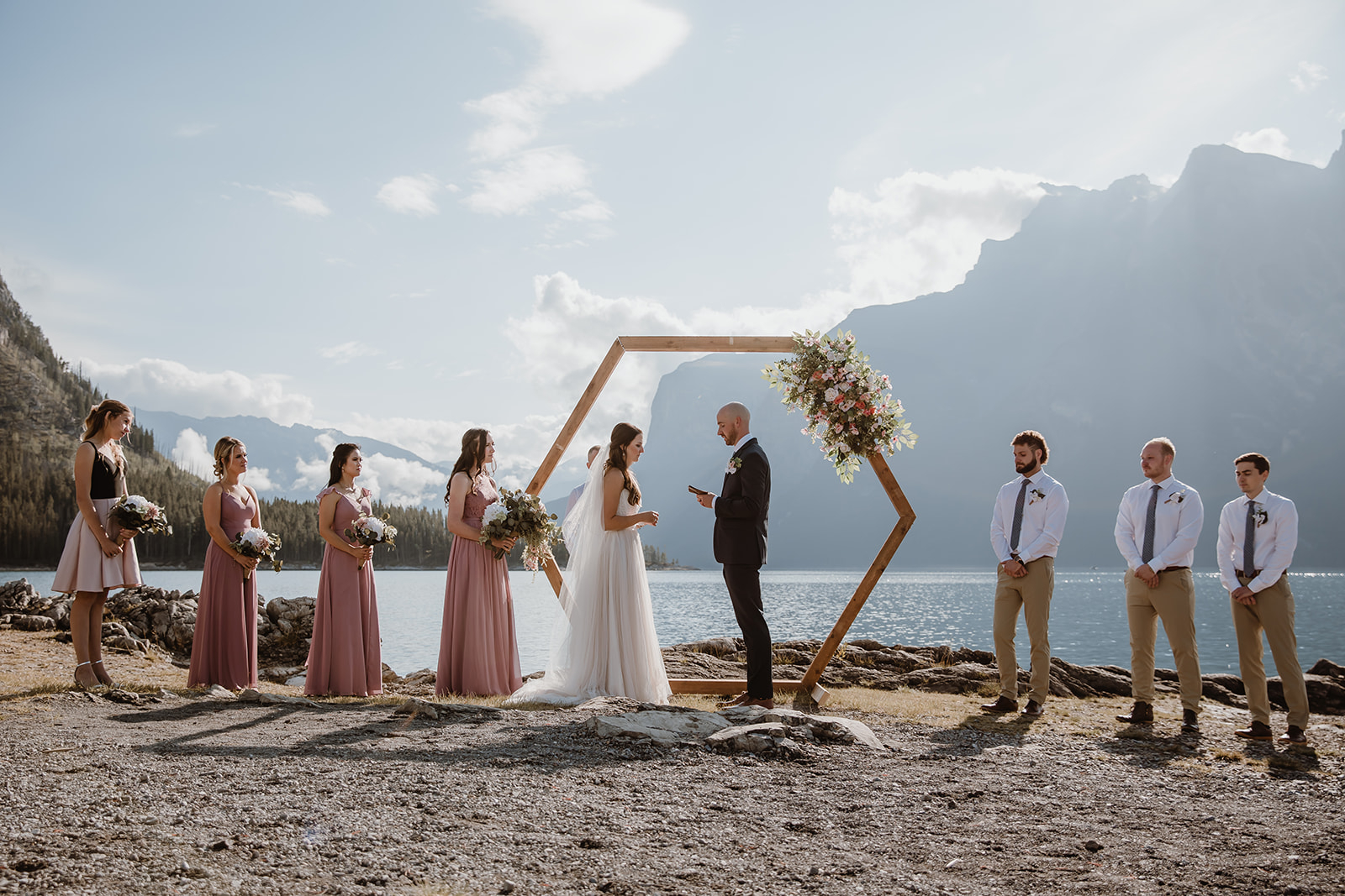 A lakeside ceremony unfolds beneath mountain peaks, with soft sunlight, floral accents, and joyful loved ones gathered.