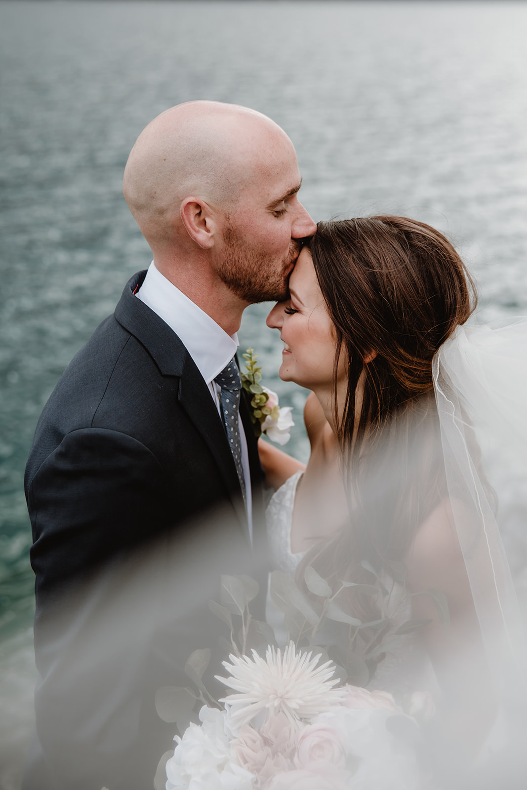 lake-Minnewanka-wedding A tender moment as the groom kisses the brideโs forehead, framed by soft florals and lake water.