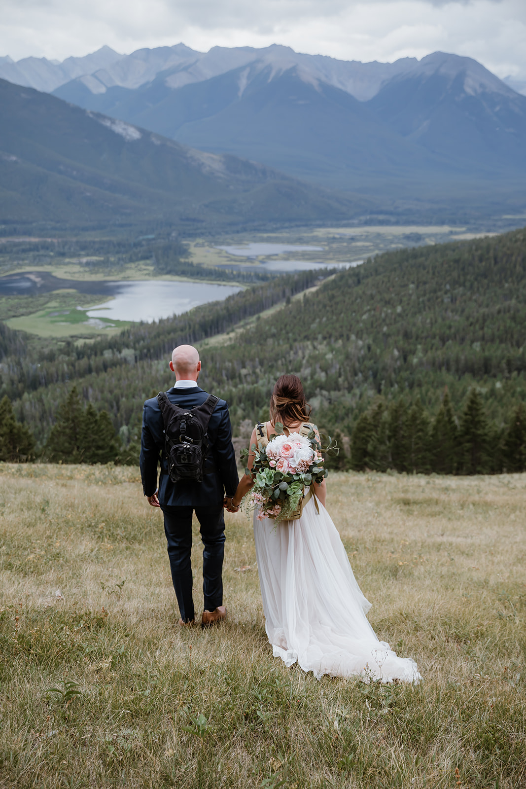 lake-Minnewanka-wedding A couple holds hands, looking out over a vast valley of lakes and forested mountains, bouquet in her backpack