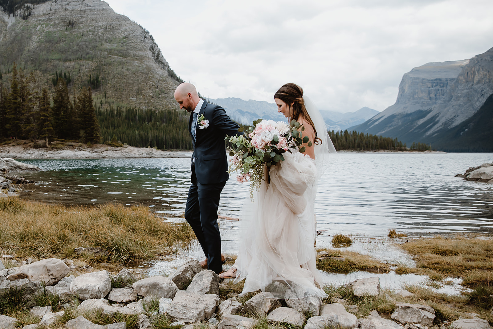 lake-Minnewanka-wedding A couple walks carefully along the rocky lakeshore, surrounded by mountains and holding a lush bouquet during their Lake Minnewanka Wedding