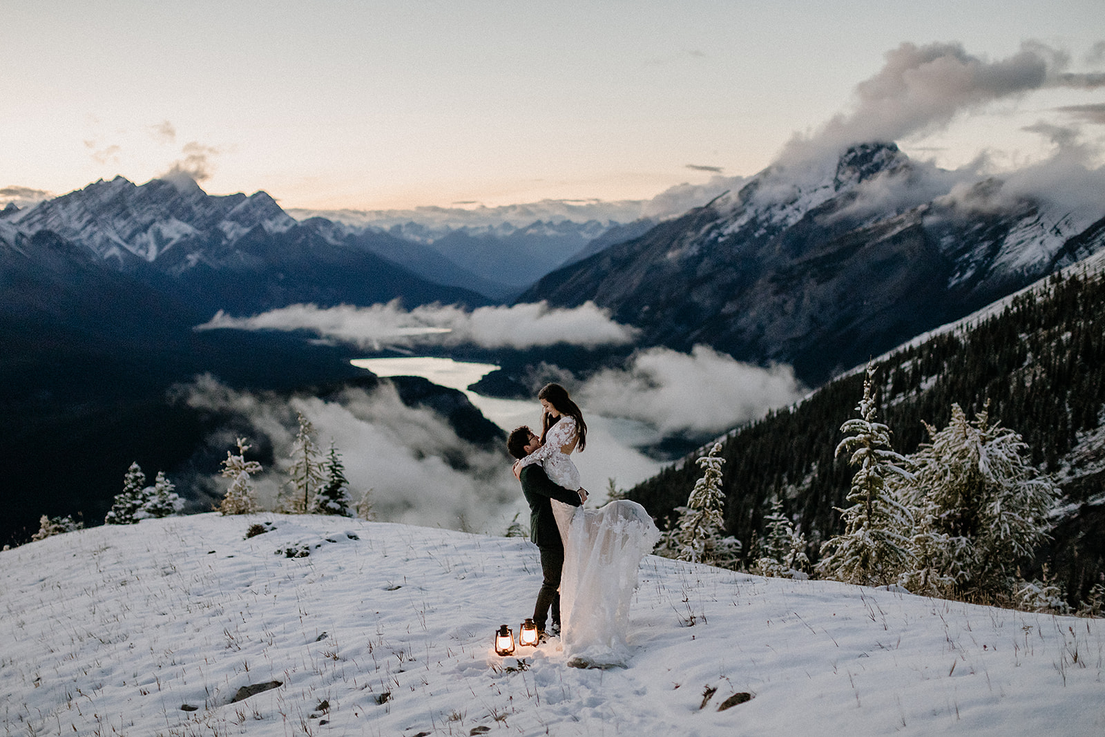 Groom lifted his bride during sunset on a snowy mountain ledge, with clouds floating over the valley and a winding lake far below.