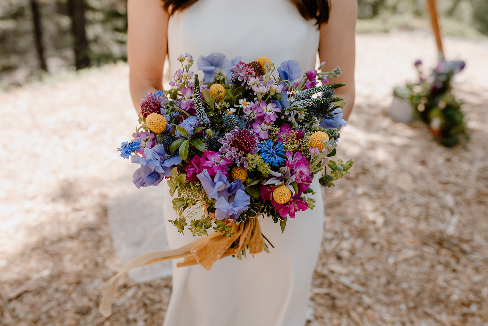 Close-up of a colorful wildflower bouquet held by a bride, featuring purple, blue, yellow, and pink blooms with greenery and a velvet ribbon.

