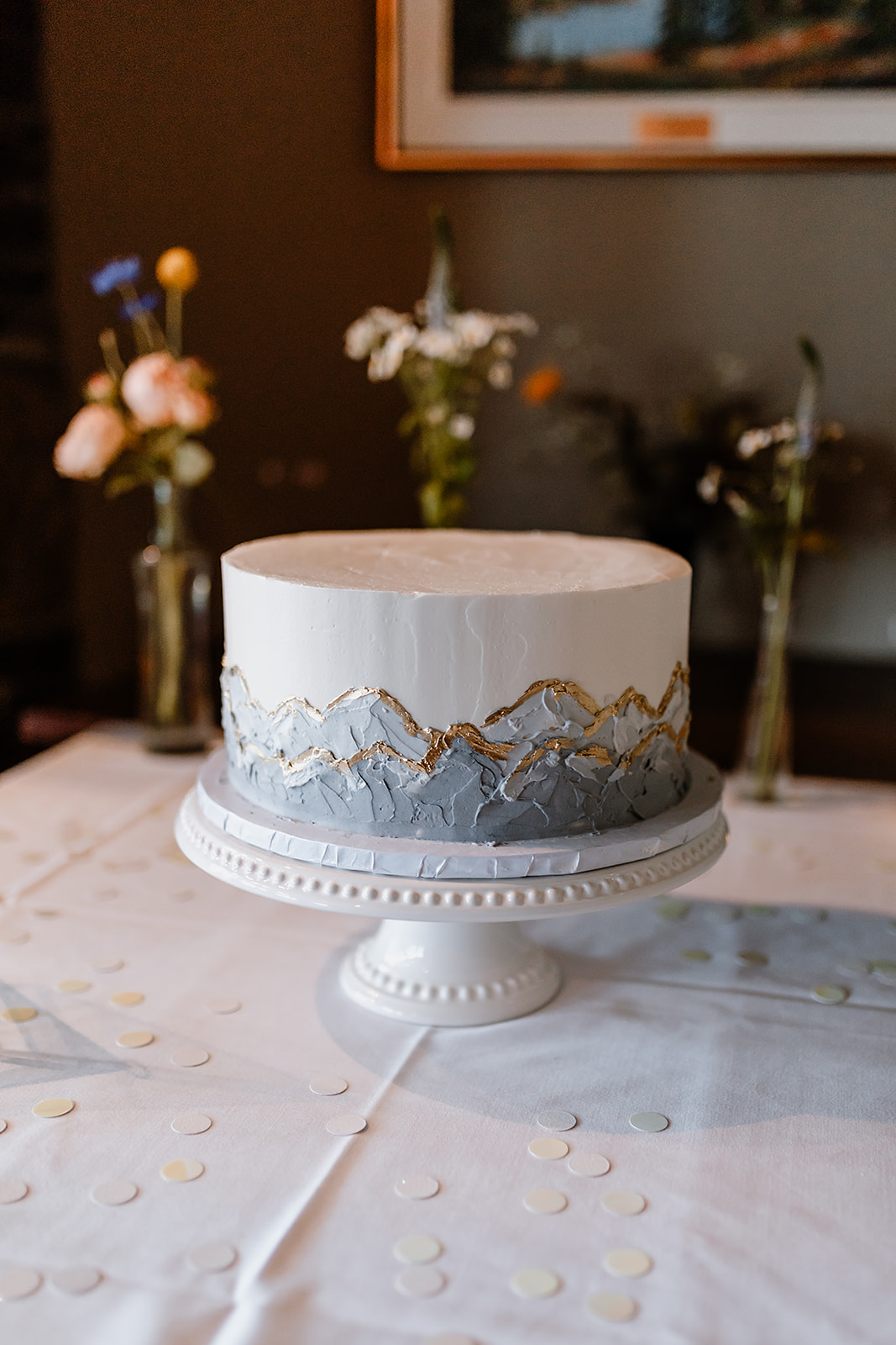 Minimalist white wedding cake decorated with hand-painted mountain details and gold accents, displayed on a white cake stand with soft floral decor in the background.