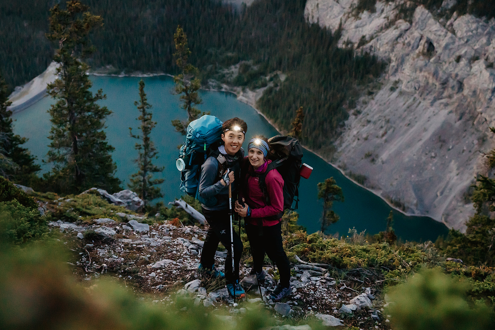 Couple smiling with headlamps and backpacks during their early morning hiking elopement in Banff, overlooking a turquoise alpine lake.