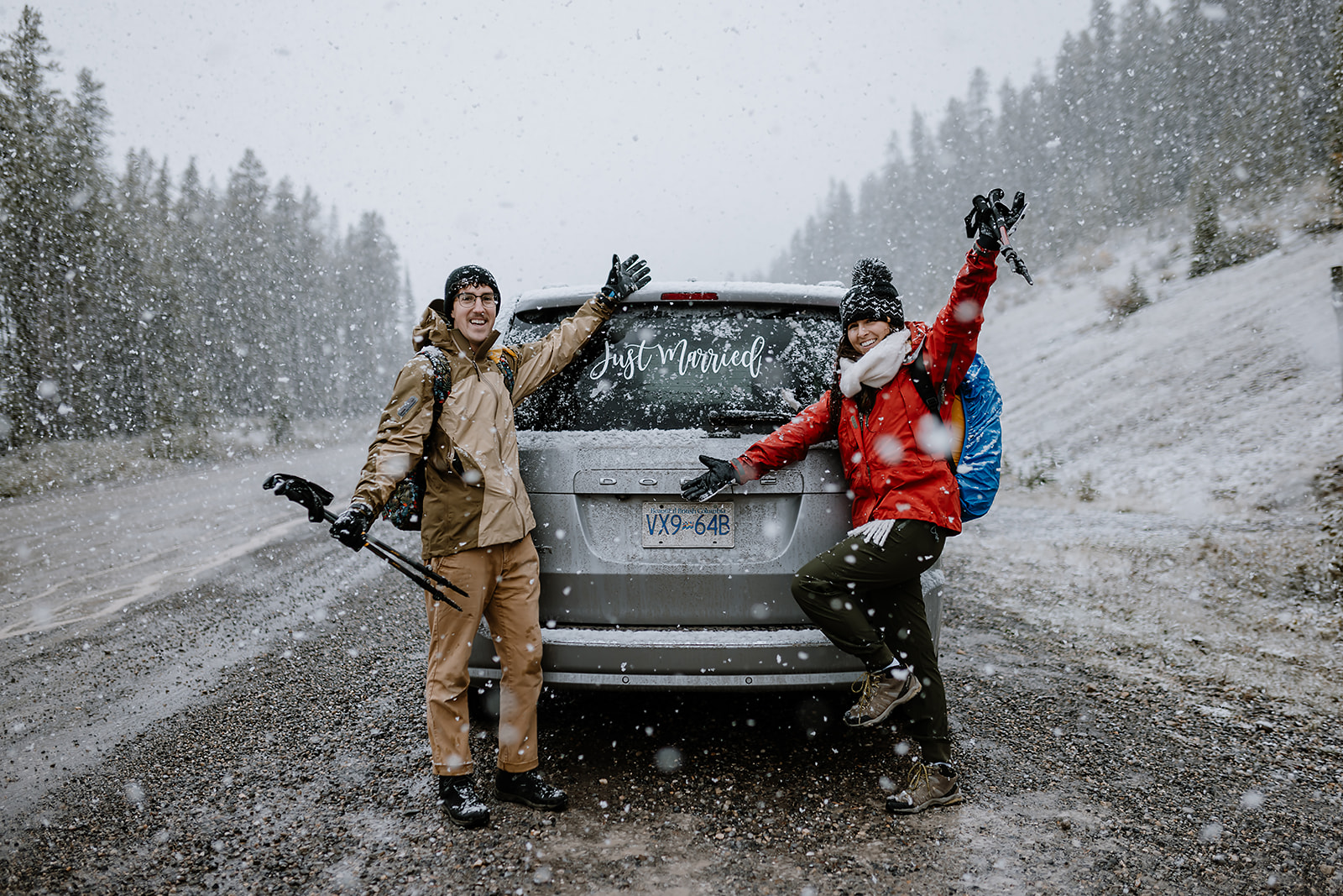 Couple celebrating next to their vehicle in the snow, holding hands and gear with joyful expressions on a wintry mountain road.