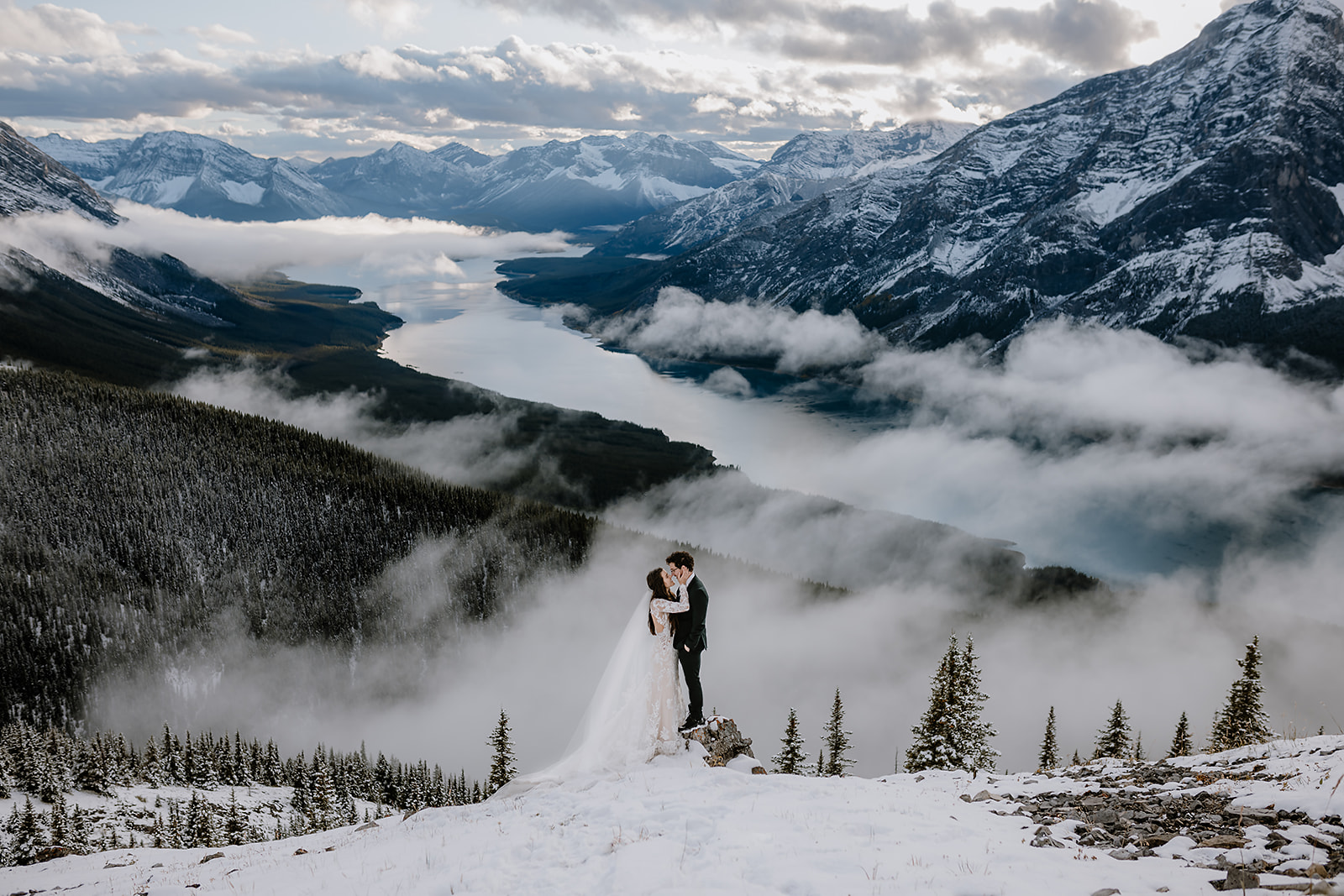 Bride and groom sharing a kiss on a snowy mountain ledge, with clouds floating over the valley and a winding lake far below.