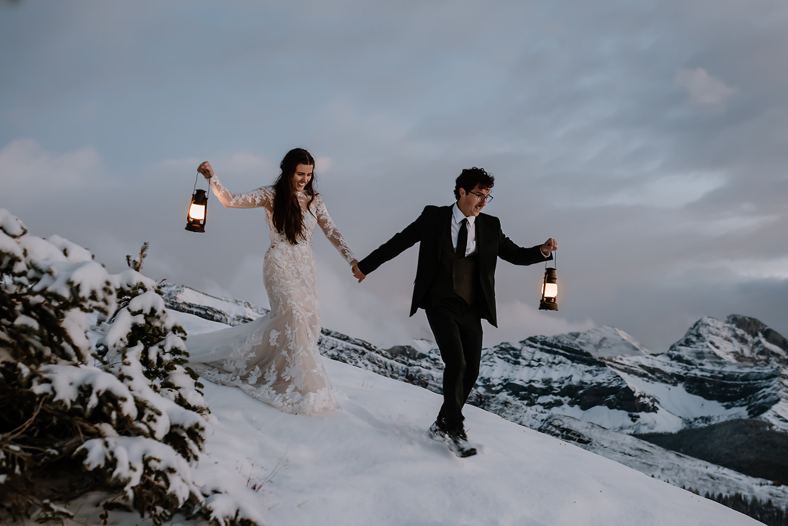 Bride and groom holding hands and walking through snow, with the groom carrying a lantern and mountains in the background.

