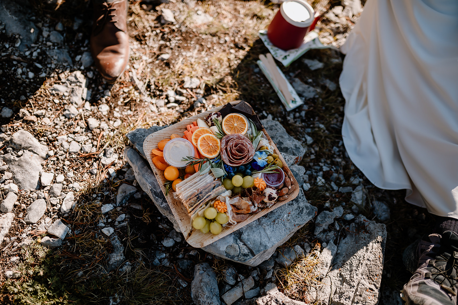 Close-up of a mountain picnic spread on rocky ground during a hiking elopement in Banff, with wedding attire just visible in the frame.


