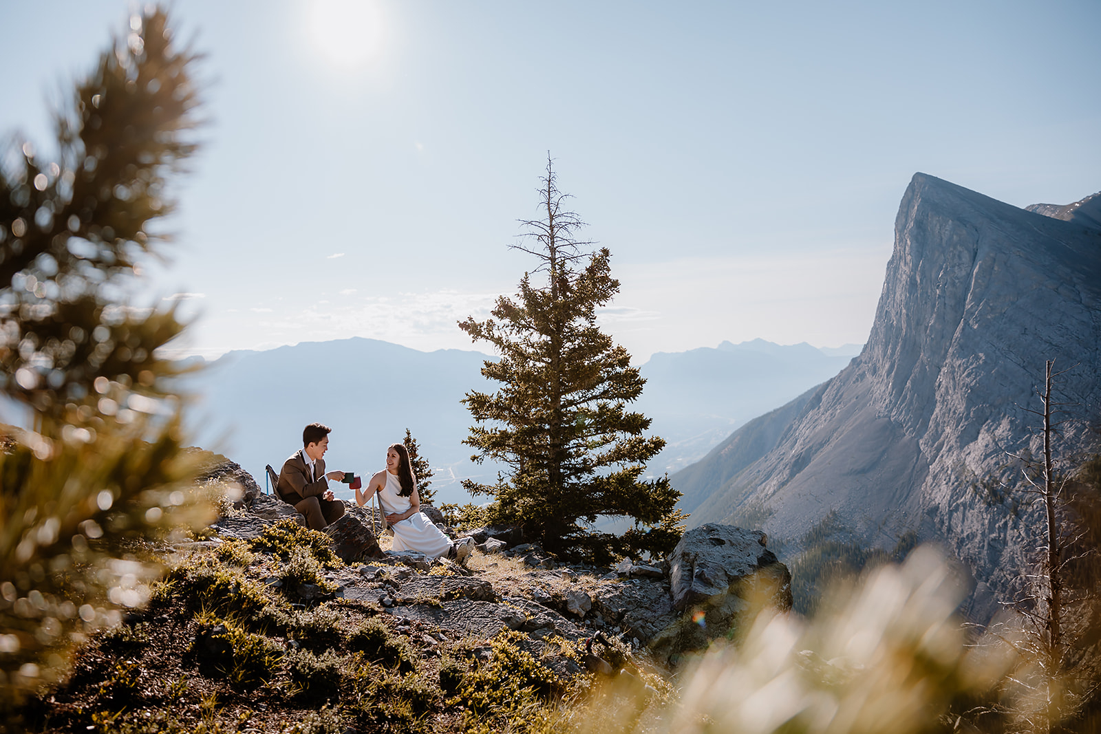 Couple sharing a quiet moment during their hiking elopement in Banff, seated on a mountain ledge with coffee and views of rugged peaks.