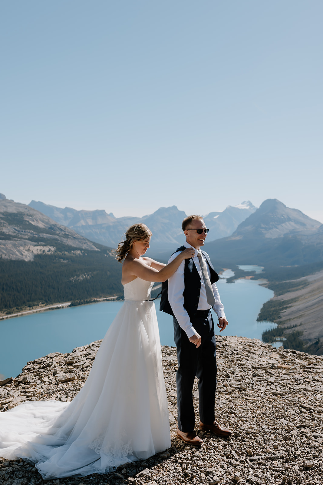 Bride helps her partner with their jacket during a hiking elopement in Banff, standing on a cliffside with Peyto Lake and mountain peaks in the background.